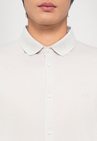 Polo-skjorte i lys beige med en teksturert hvit krage, med fem knapper og en liten brodert logo på brystet.