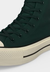 Σκούρο πράσινο sneaker ψηλής κοψής με μαύρα μεταλλικά κορδόνια, ταιριαστά πράσινα κορδόνια, λευκή λαστιχένια άκρη μπροστά, και παχιά λευκή σόλα με μαύρη ρίγα.