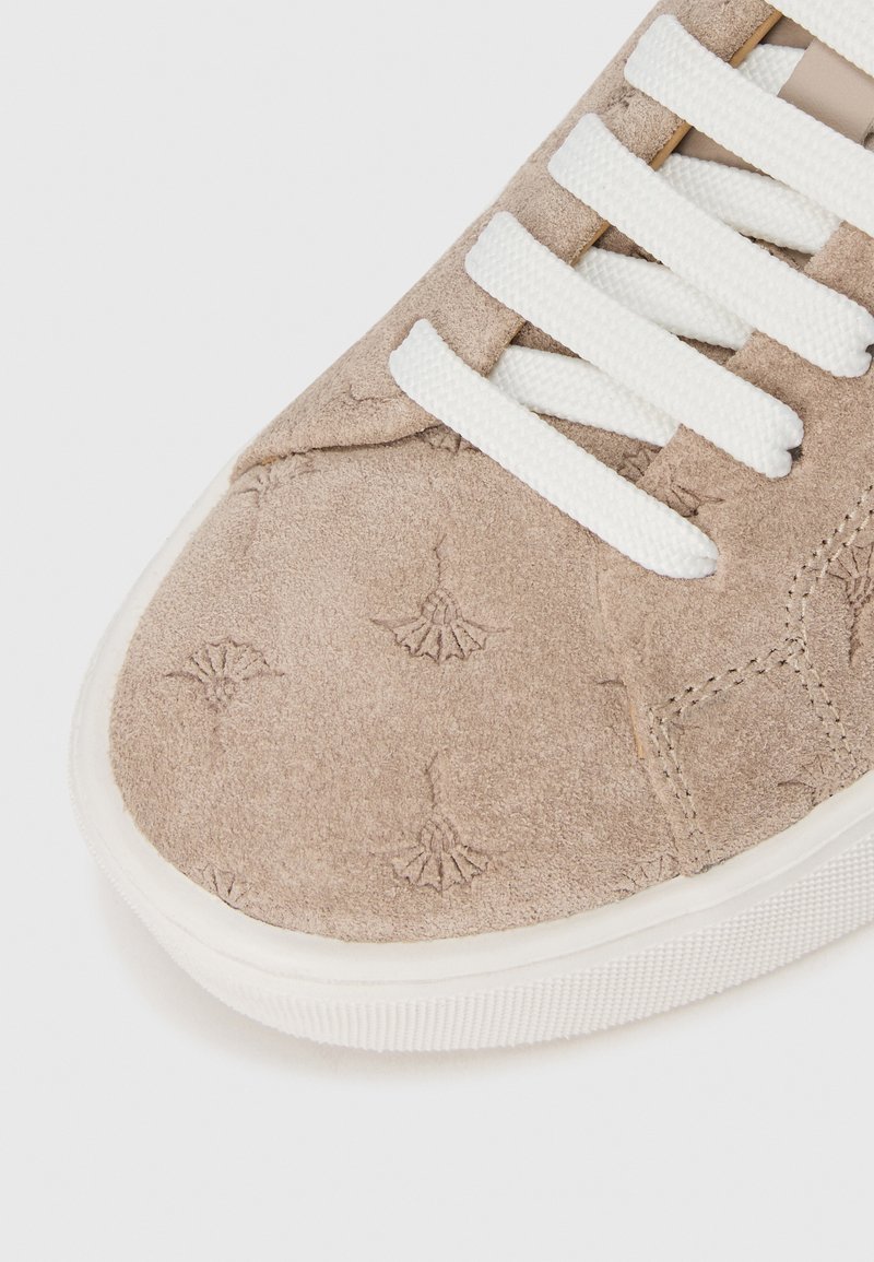 Beige Wildlederschuh mit weißen Schnürsenkeln, der ein dezentes Blumenmuster aufweist. Gerundete Zehenpartie und strukturierte weiße Gummisohle.