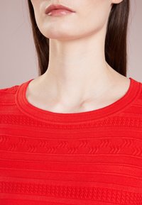 Visage inférieur et cou d'une femme portant un haut en maille rouge texturée avec un col rond sur un fond rose doux.