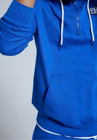 Sininen vetoketjullinen collegepaita, jossa valkoiset kiristysnauhat, kengurutasku ja resorireunukset. Pehmeä pinta, yksivärinen ilman kuvioita.