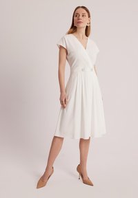 Biała sukienka wrap z krótkimi rękawami, dekoltem w kształcie litery V, z wcięciem w talii podkreślonym dwoma guzikami oraz rozkloszowanym dołem. Gładka tekstura materiału.