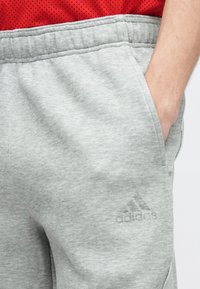 Άτομο που φοράει ανοιχτό γκρι φόρμα Adidas με το χέρι στην τσέπη και κόκκινο πουκάμισο με διάτρητο σχέδιο, μαζεμένο μέσα από το παντελόνι.