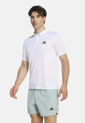 Junger Mann trägt weißes Polohemd mit kurzen Ärmeln und hellblaue Shorts, beide mit schwarzem Adidas-Logo, steht vor neutralem Hintergrund.