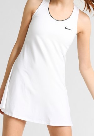 Vrouw draagt een witte mouwloze Nike-tennisjurk met zwarte biezen rond de halslijn tegen een effen achtergrond.