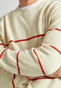 Cremefarbener Strickpullover mit roten horizontalen Streifen, gerippter Textur und Rundhalsausschnitt. Das Material wirkt weich und leicht grob.