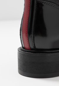 Botín de cuero negro con un acabado brillante, que presenta una franja vertical roja y un grueso tacón de madera negra. Costuras detalladas visibles.