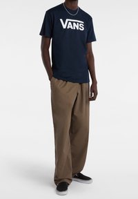 Marineblauwe Vans-t-shirt met witte logo, gecombineerd met beige wijde broek en zwarte skate schoenen. Geweven stof; relaxed fit.