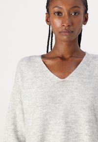 Pull en V gris clair en tissu doux et texturé ; présente une coupe décontractée et des accents côtelés au niveau du col et des poignets.