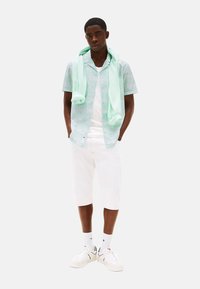 Camisa de manga corta de color verde menta con estampado, pantalones cortos blancos y zapatillas blancas, con un suéter a juego de color menta sobre los hombros.
