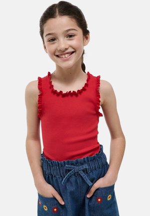 Fille souriante aux cheveux foncés tressés, portant un haut rouge sans manches à volants et un jean bleu avec des fleurs brodées, mains dans les poches.