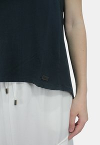 Top senza maniche di colore blu navy con una vestibilità ampia, aperture laterali e dettagli in metallo argentato. Il tessuto è liscio e presenta una leggera lucentezza. Tag del marchio sull'orlo.