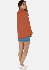 Rost-orange Langarmshirt mit Kragen, geknöpften Manschetten und abgerundetem Saum, kombiniert mit einem Jeansrock und weißen Slip-On-Schuhen.