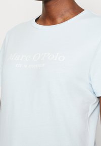 Maglietta in cotone blu chiaro con colletto a girocollo. Presenta un logo stampato bianco "Marc O'Polo" e "EST. IN STOCKHOLM" sul fronte. Maniche corte.
