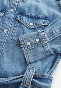 Gros plan sur une veste en denim bleu montrant une poche avant avec rabat à bouton, un poignet avec deux boutons et une passant de ceinture avec une couture contrastée.