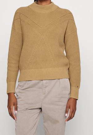 Osoba ubrana w sweter z grubego, camelowego dzianego materiału z wzorem w jodełkę oraz jasnobeżowe, wysokowstanowe spodnie, stojąca na tle jednolitego tła.