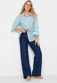 Camicia azzurro chiaro con dettagli di occhiello, abbottonata, sovrapposta a jeans a gamba larga blu scuro. Modella in piedi con sandali, mani sui fianchi.