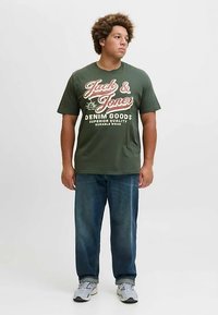 Žalia medvilninė marškinėliai su baltais ir raudonais tekstiniais grafikos elementais. Derinama su mėlynais džinsais, dėvima su pilkais sneakeriais.