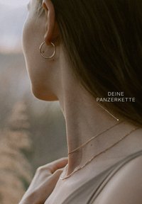 Goldfarbene Creolen und geschichtete schlanke Ketten liegen auf einer nackten Schulter. Der Hintergrund ist sanft unscharf mit natürlichen Tönen.