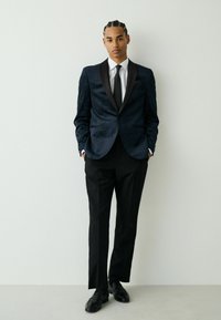 Sacou din catifea bleumarin cu rever din satin, cămașă albă, cravată neagră cu dungi și pantaloni negri. Design clasic cu croială ajustată.