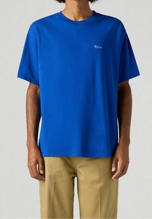 Homme portant un t-shirt uni bleu roi avec "Paris" brodé sur la poitrine, assorti à un pantalon beige, debout devant un fond uni.