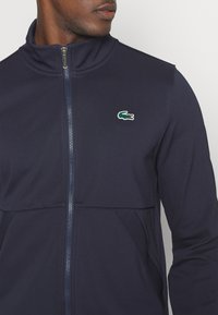 Marineblaue Zip-Jacke mit Stehkragen, strukturiertem Stoff und kleinem grünen Krokodil-Logo auf der linken Brust. Mit seitlichen Taschen.