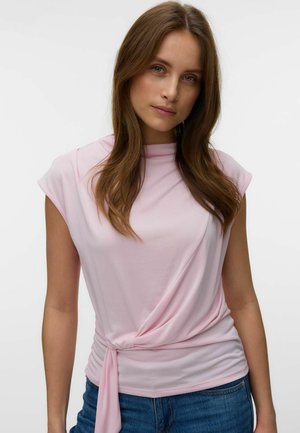 Jeune femme aux longs cheveux bruns portant un haut sans manches rose pâle avec un nœud sur le côté et un jean bleu sur un fond uni.