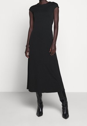 Femme portant une robe noire à manches courtes, longue jusqu'aux chevilles, associée à des bottes en cuir noires montant jusqu'aux genoux, debout devant un fond blanc uni.