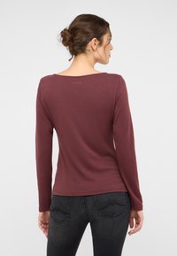 Bordo spalvos ilgomis rankovėmis marškiniai iš minkštos, lygios medžiagos su plačia iškirpte ir minimalia prekės ženklo žyme nugaroje, derinami su tamsiomis džinsomis.