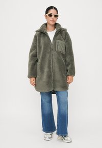 Olivgrön fleecemantel med huva, dragkedja och bröstficka. Bärs över en vit topp och vida jeans med en texturerad yta.