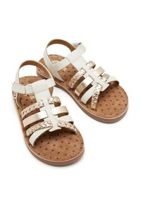 Sandalias de cuero blanco y dorado metálico con correas trenzadas, suela plana e interior marrón con un patrón de estrellas. Incluye una correa ajustable para el tobillo.