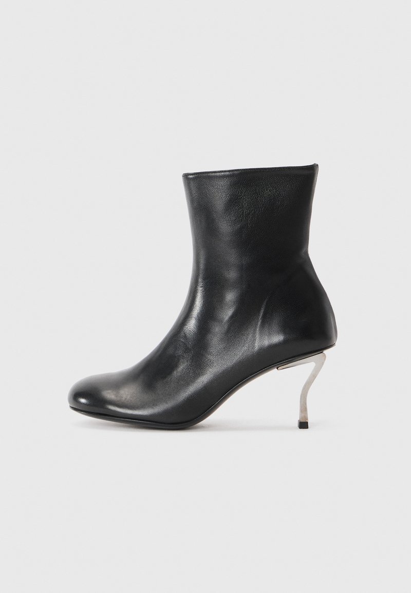Bota de tornozelo em couro preto com um salto agulha prateado curvo sobre um fundo branco.