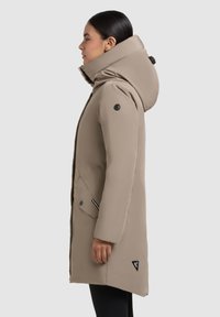 Woman wearing a beige knee-length winter coat with high collar and large hood, standing in side profile against a plain background.