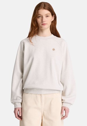 Kvinde med langt rødt hår iført en lys beige sweatshirt og cremefarvede kordfløjlsbukser mod en ensfarvet hvid baggrund.