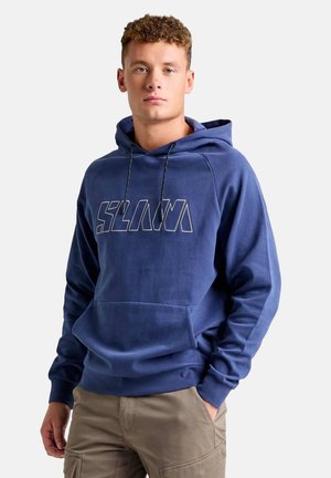 Junger Mann trägt ein blaues Kapuzensweatshirt mit Kängurutasche vorne und beige Cargohosen, steht vor einem schlichten hellen Hintergrund.