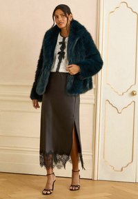 Zilgli zaļi mākslīgie kažokādas svārki, balta mežģīņu blūze ar melnu loku, glīti melni satīna svārki ar mežģīņu maliņu un melnas siksniņu kurpes.