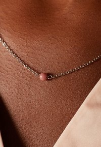 Collier en chaîne argentée avec une perle ronde rose, reposant sur une peau brune texturée avec un tissu de couleur claire en arrière-plan.