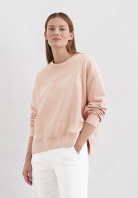 Hellrosa Sweatshirt mit Rundhalsausschnitt und gerippten Bündchen. Verfügt über ein embossed Logo auf der Vorderseite und einen lässigen Schnitt. Kombiniert mit weißen Hosen.