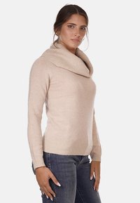 Maglione beige chiaro con collo a cowl realizzato in materiale morbido e testurizzato, con maniche lunghe e silhouette aderente. Indossato con jeans scuri.