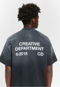 Schwarzes, oversized T-Shirt mit Rundhalsausschnitt, auf dem Rücken mit weißem Text: "CREATIVE DEPARTMENT 2018 CD" in fetter Schrift. Weiche Stofftextur.
