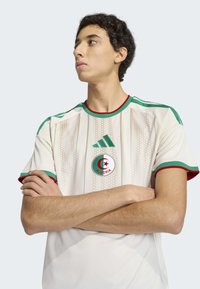 Wit voetbalshirt met rode en groene accenten, voorzien van een ronde hals, horizontale strepen en het Algerije-logo op de borst.