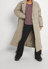 Abrigo largo de piel beige acolchado, top de estampado floral, pantalones negros acampanados con detalle de cordones en los dobladillos y zapatos negros de punta redonda.