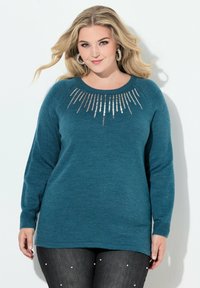 Türkisfarbener Pullover mit langen Ärmeln, gerippten Bündchen und rundem Halsausschnitt. Mit silberfarbigen Paillettenverzierungen über der Brust dekoriert.