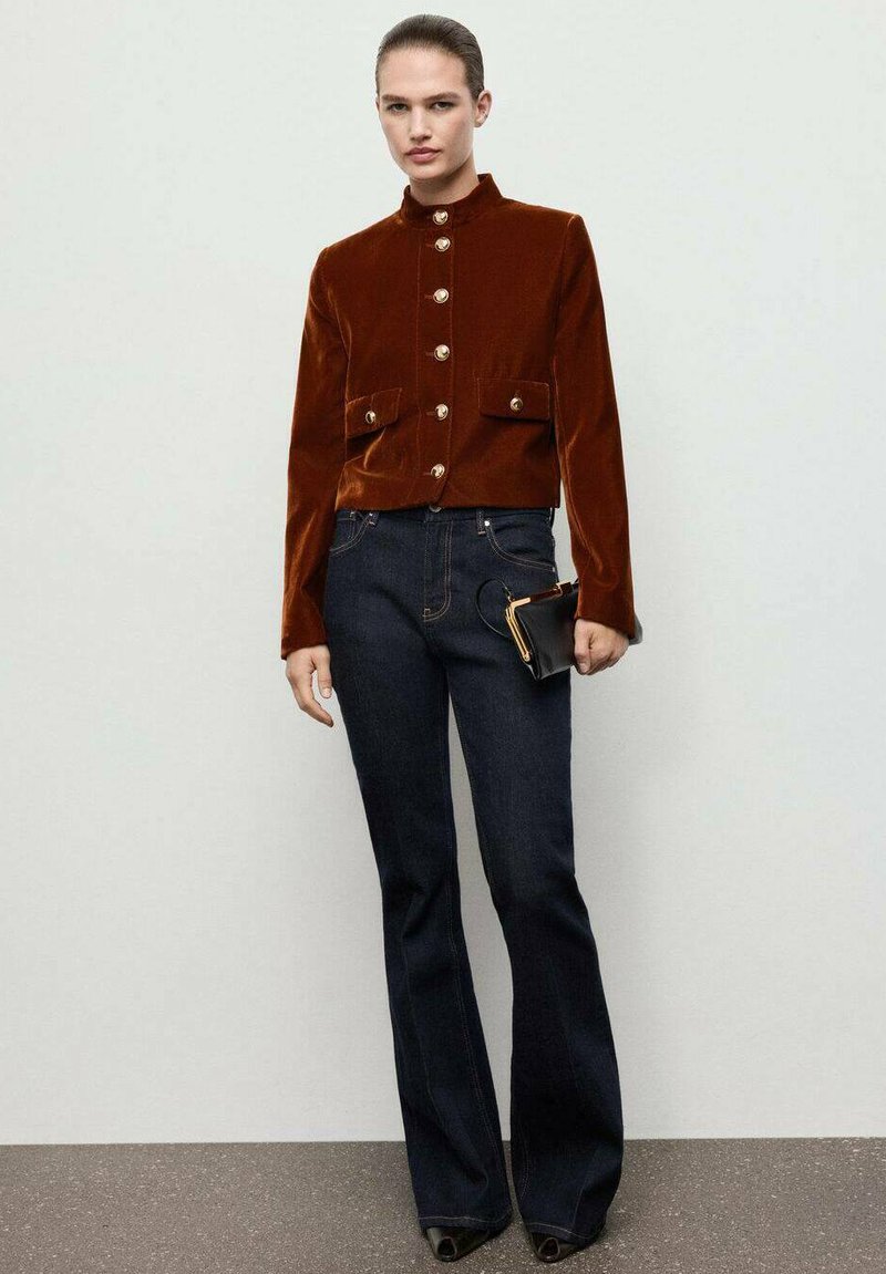 Giacca corta in velluto marrone con bottoni dorati e due tasche frontali, abbinata a jeans svasati blu scuro e una piccola pochette nera.
