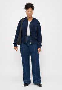 Ναυτική πλεχτή ζακέτα με φερμουάρ και υφή, συνδυασμένη με ψηλόμεσα σκούρα τζιν και μαύρες παντόφλες.