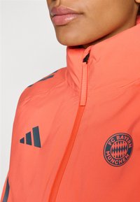 FC Bayern München jack in fel oranje. Heeft een hoge kraag, ritssluiting en donkerblauwe accenten waaronder het logo en strepen.