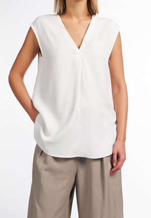 Blouse sans manches en tissu crème clair, avec un décolleté en V, présentant une coupe décontractée, une texture lisse et un pli central subtil. Associée à un pantalon beige.