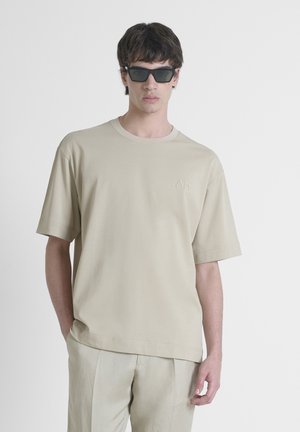 T-shirt beige oversize à manches courtes, col rond, avec un détail de logo subtil sur la poitrine, associé à un pantalon de couleur claire.