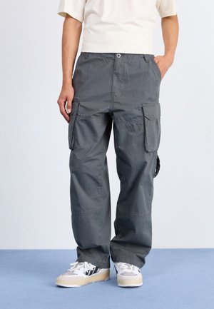 Persona con pantalones cargo grises y zapatillas blancas, de pie con una mano en el bolsillo contra un fondo liso.