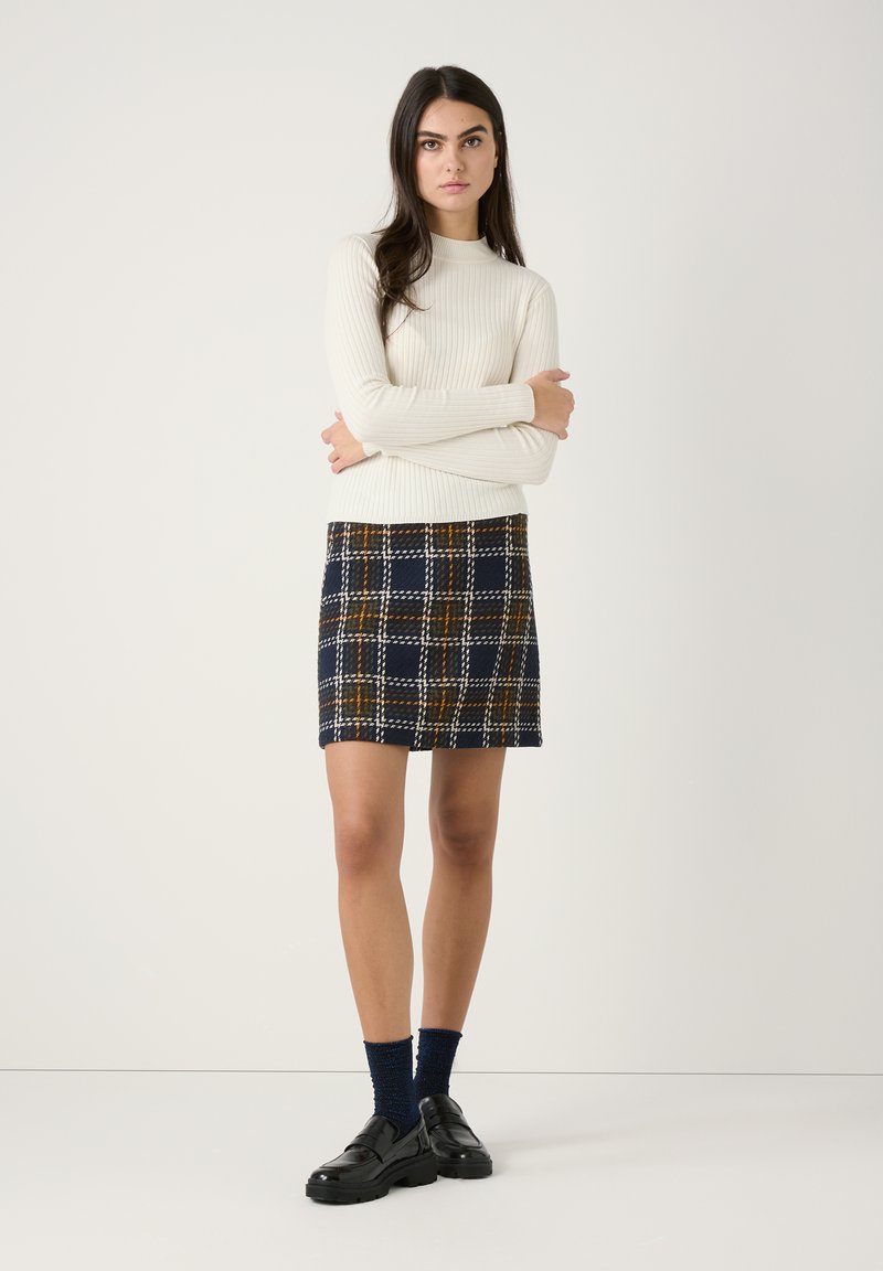 Knit sweater in cream with ribbed texture, paired with a navy plaid skirt featuring yellow and white accents. Black shoes and blue socks.